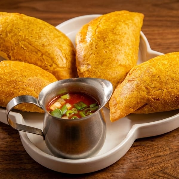 Four golden-brown empanadas on a white plate with a silver pitcher of red sauce and green onions.