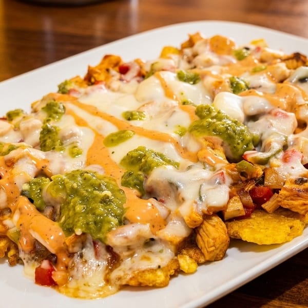 A white plate holds a dish of plantain nachos topped with chicken, melted cheese, green salsa, and pink sauce.