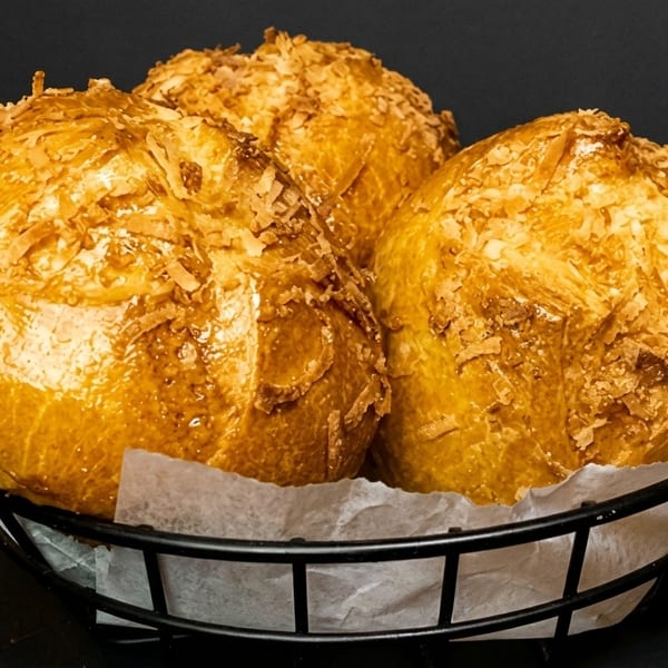 Three golden-brown coconut buns with flaky toppings sit in a black wire basket lined with white paper.